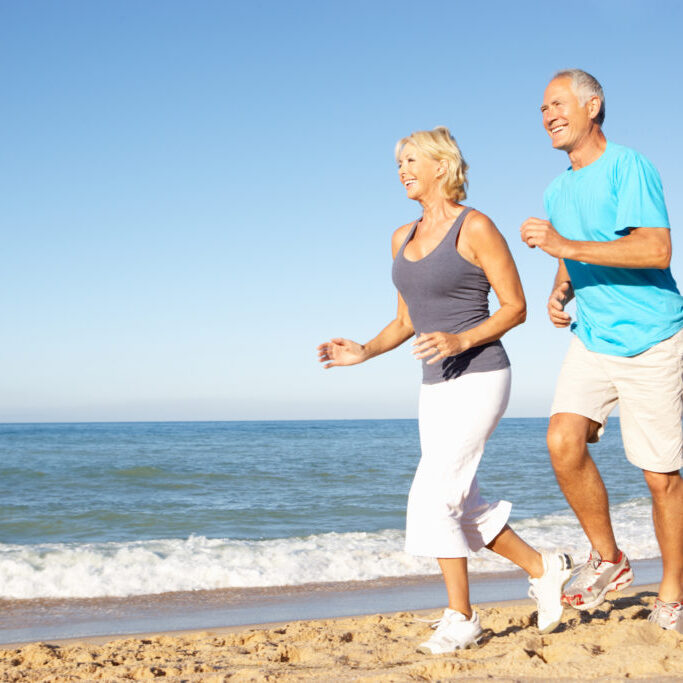 Senior Couple In Fitness Clothing Running Along Beach